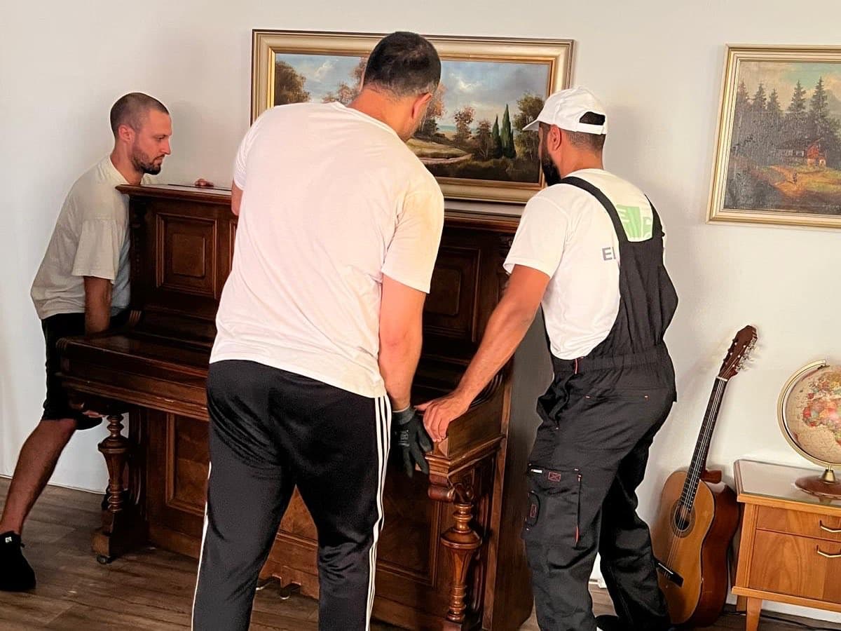Drei Mitarbeiter heben gemeinsam ein altes Klavier aus Holz an